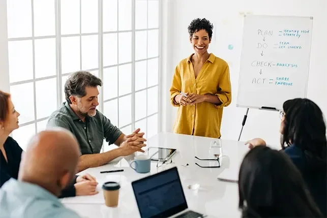 A diverse group of people attentively listens to a woman presenting during a meeting.